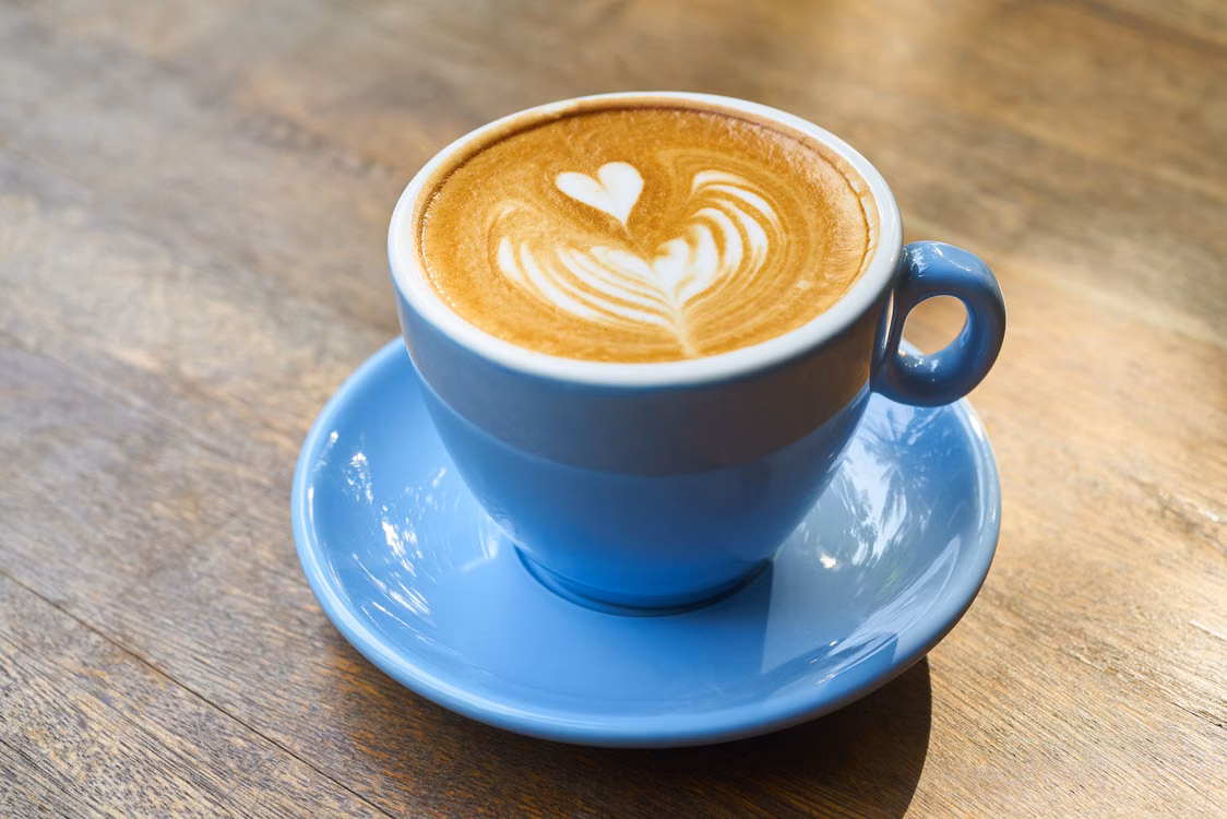 Illustrative photograph of a coffee drink with foam art in a blue cup and saucer on a wooden table.