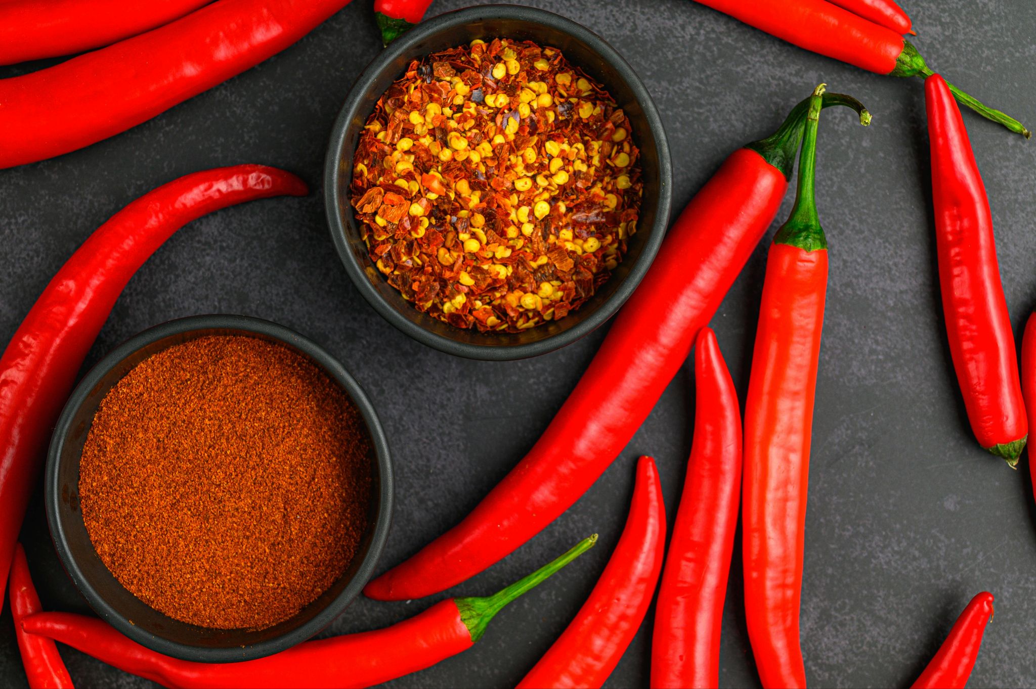 Fresh red peppers around bowls of crushed pepper flakes and ground seasoning.