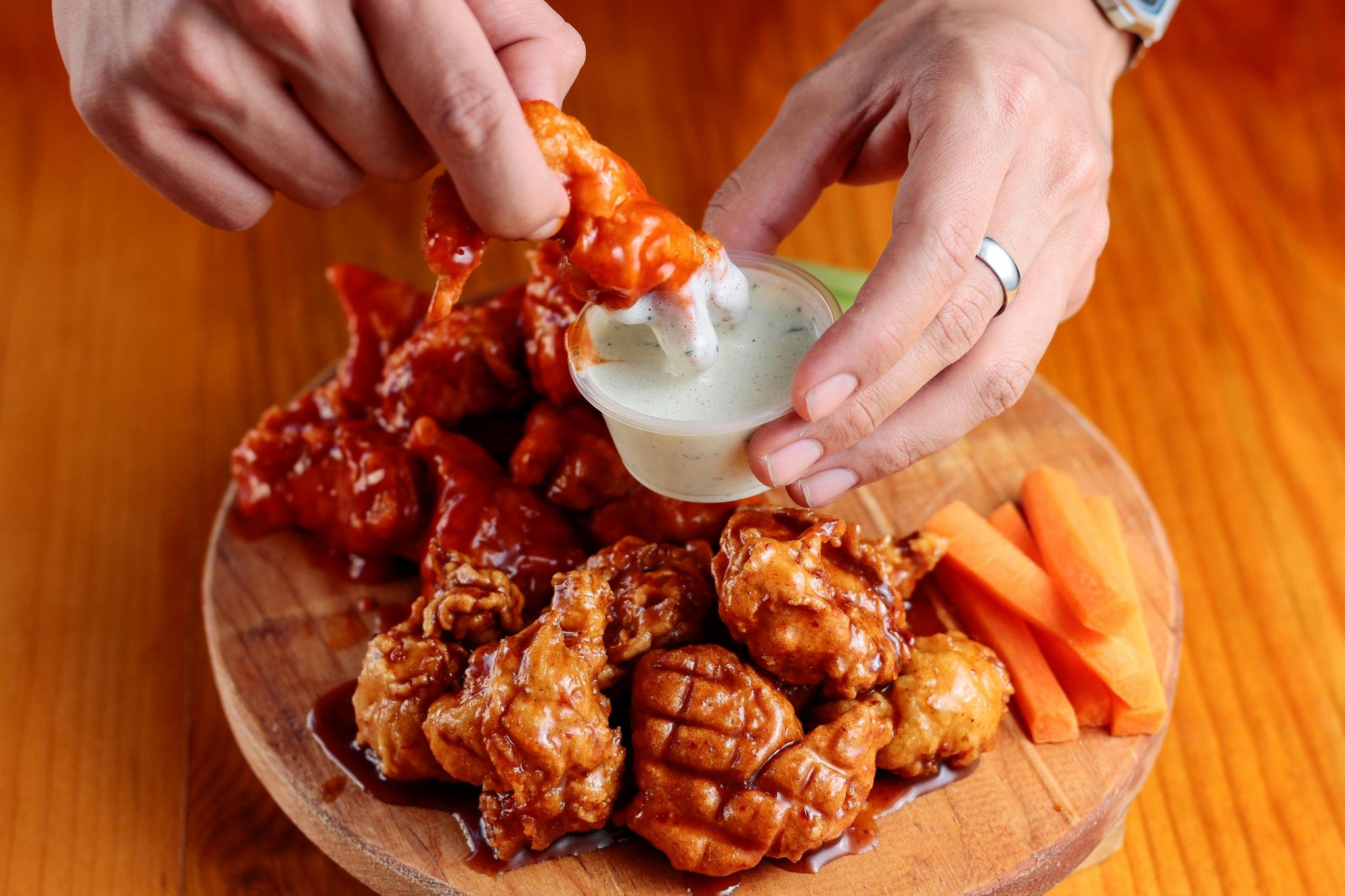 A sauced chicken wing dipped into a pale sauce beside more wings and carrot sticks.