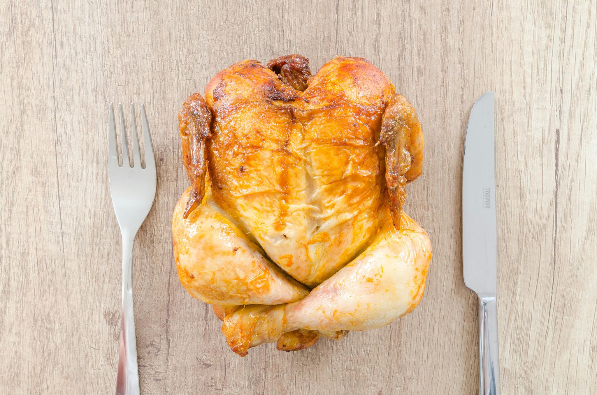 Whole roasted chicken with a fork and knife on a wooden table.