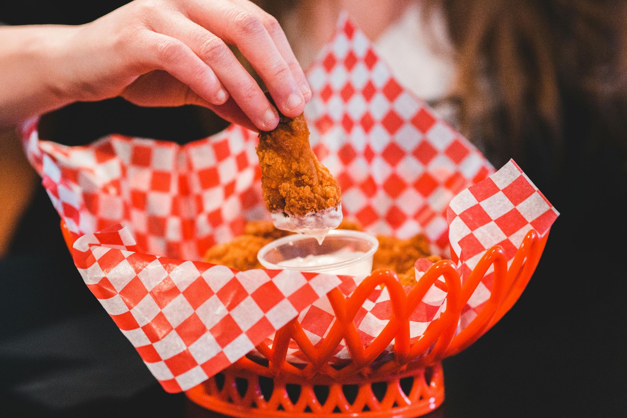 Breaded chicken dipped into a pale sauce beside a red serving basket.