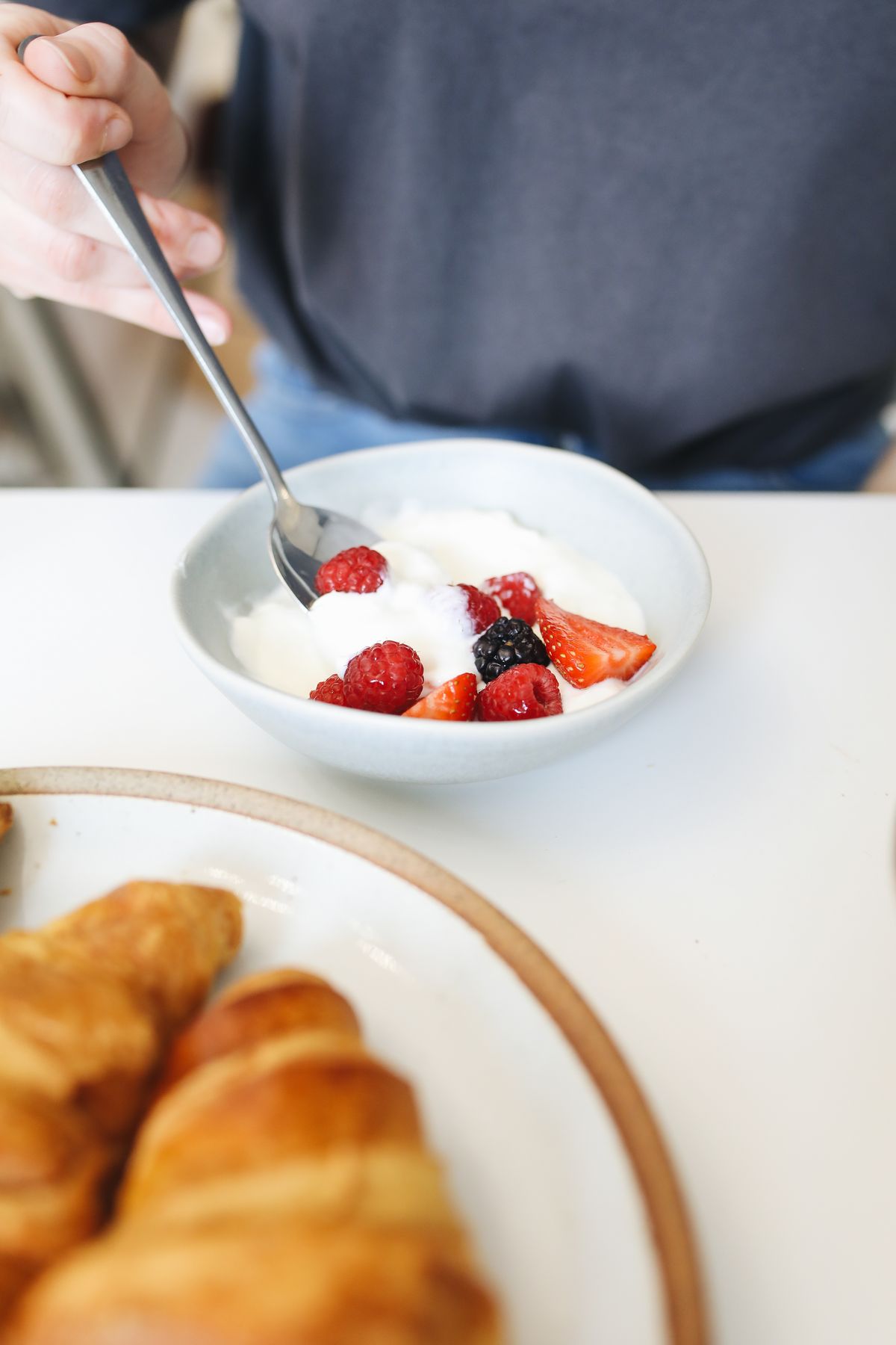 A hand holds a spoon over yogurt and berries, with croissants in the foreground.