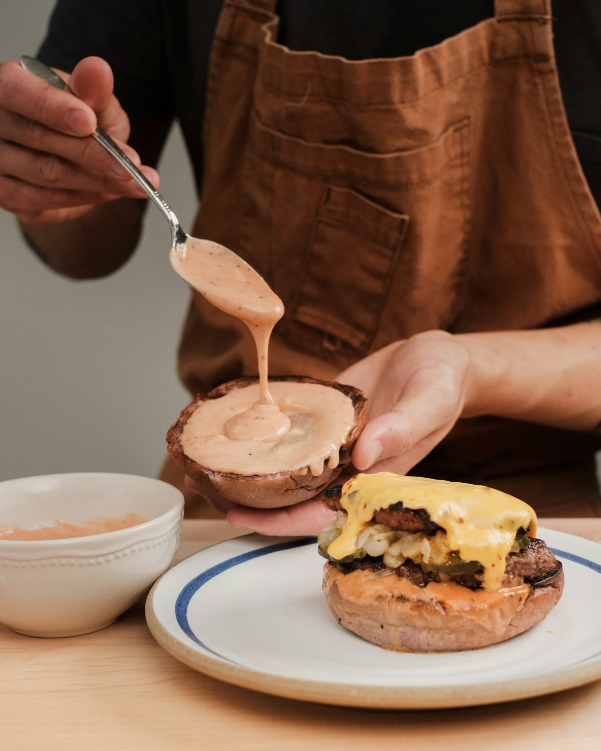 A person pouring pale pink sauce onto a bun above a cheeseburger.