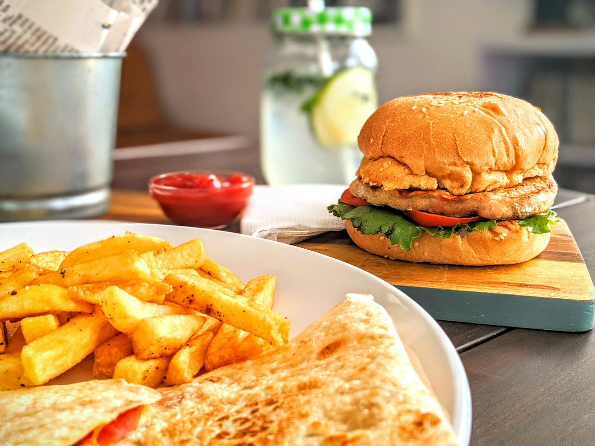 A burger on a wooden board beside fries on a white plate, with a drink behind.