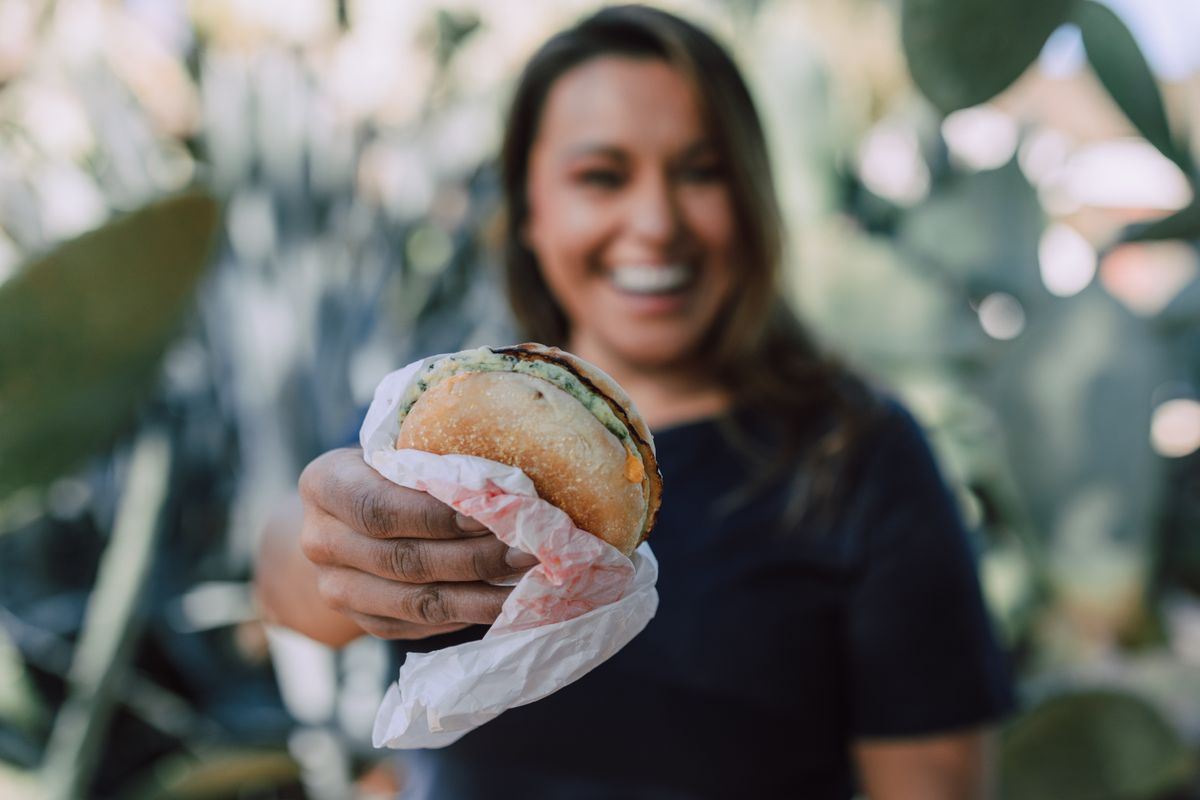 A woman holding a wrapped burger outdoors, with the burger in focus.