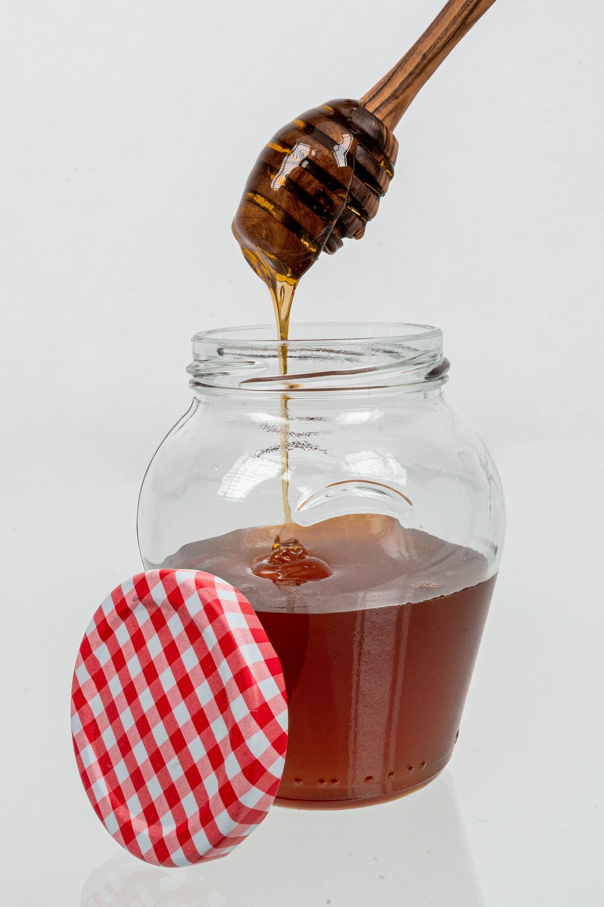 Honey dripping from a wooden dipper into an open glass jar beside a red-and-white checked lid.