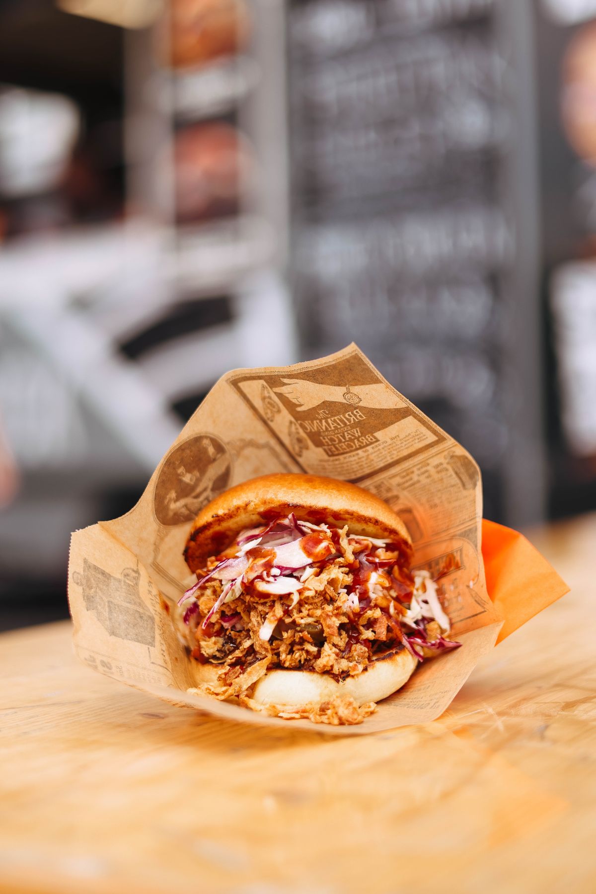 A bun filled with shredded meat, slaw and sauce rests in printed paper on a wooden counter.