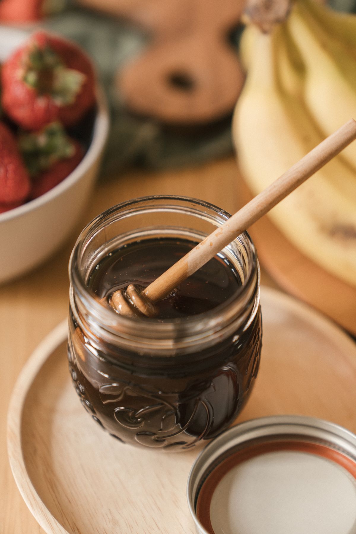 An open glass jar of dark honey with a wooden dipper on a tray beside fruit.