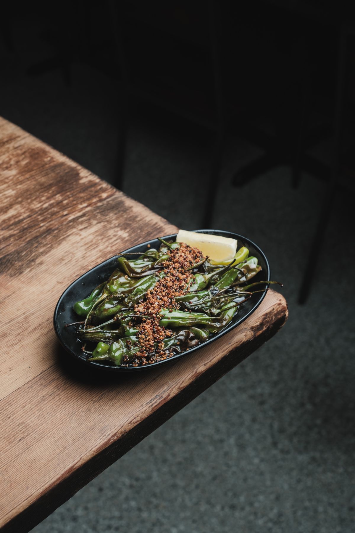 Blistered green peppers with a lemon wedge and crumb-like topping in a black dish on a wooden table.
