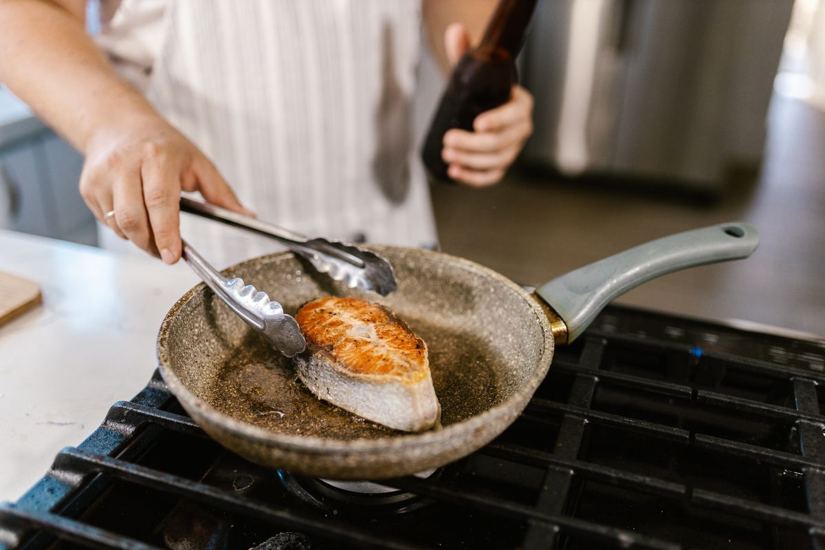 A person using metal tongs beside a browned salmon steak in a frying pan on a gas stove.