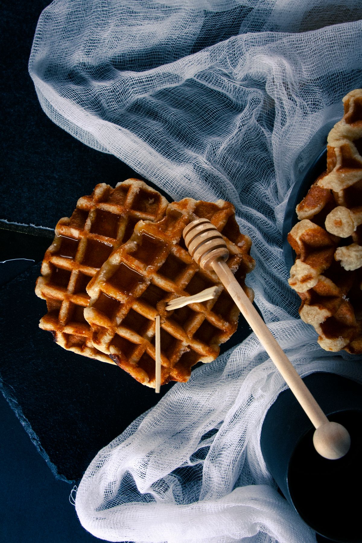Waffles and a wooden honey dipper on gauze over a dark background.