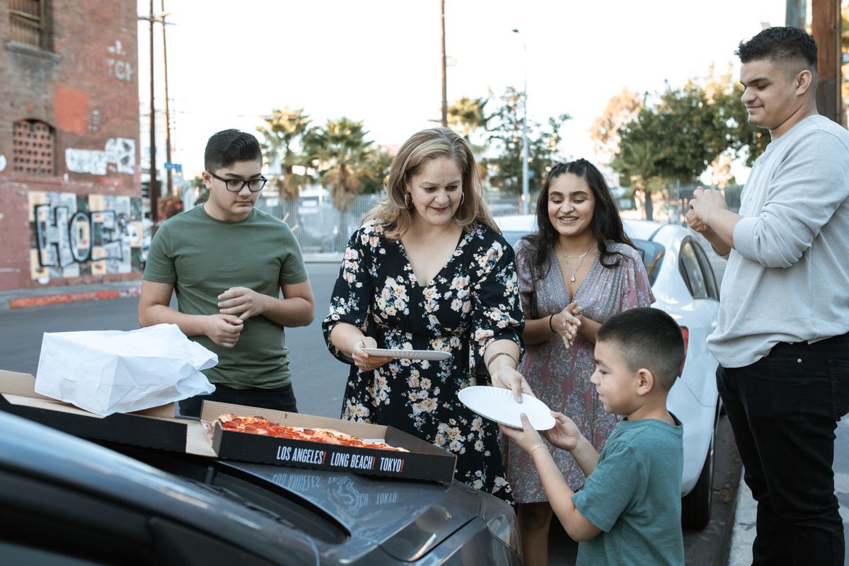 People gathered around an open pizza box beside a car.