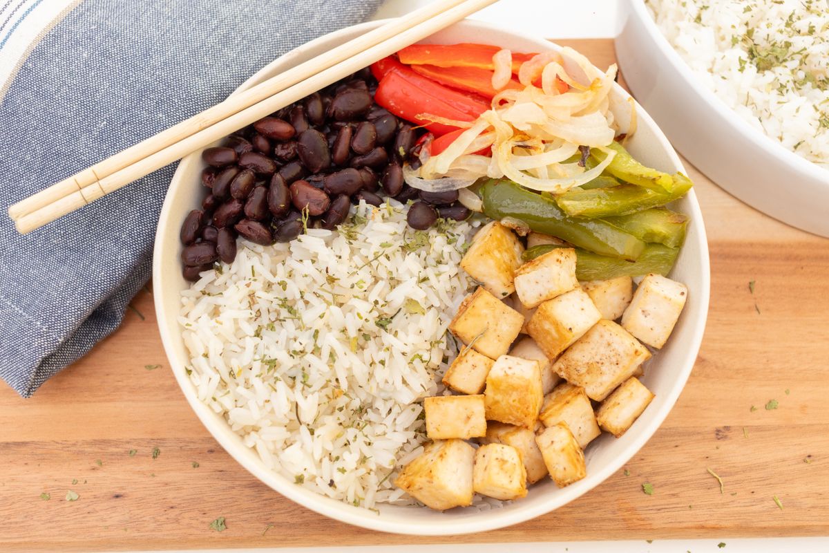 A bowl of tofu cubes, rice, black beans, peppers and onions with chopsticks resting across it.