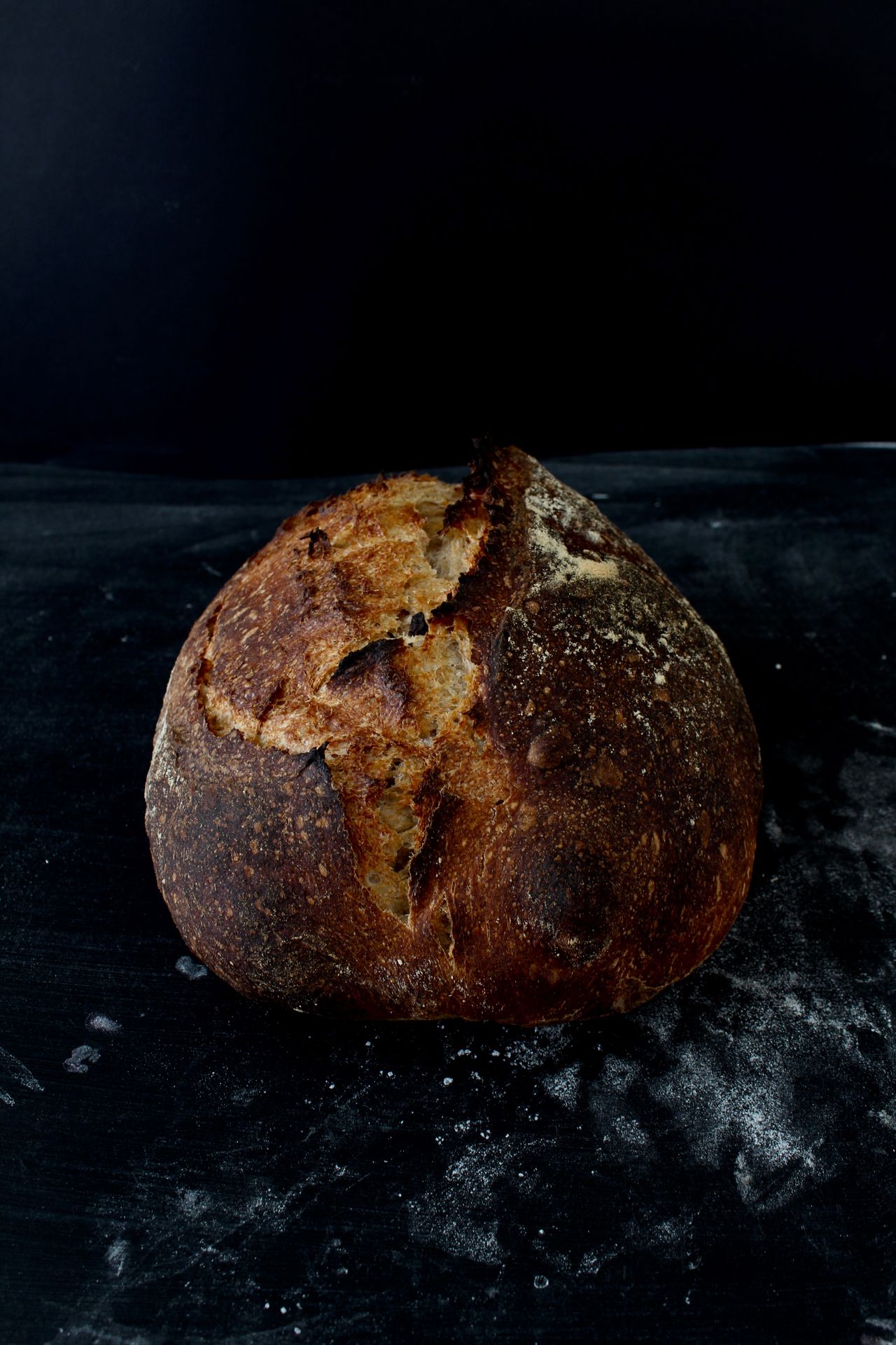 A round crusty bread loaf on a dark flour-dusted surface.