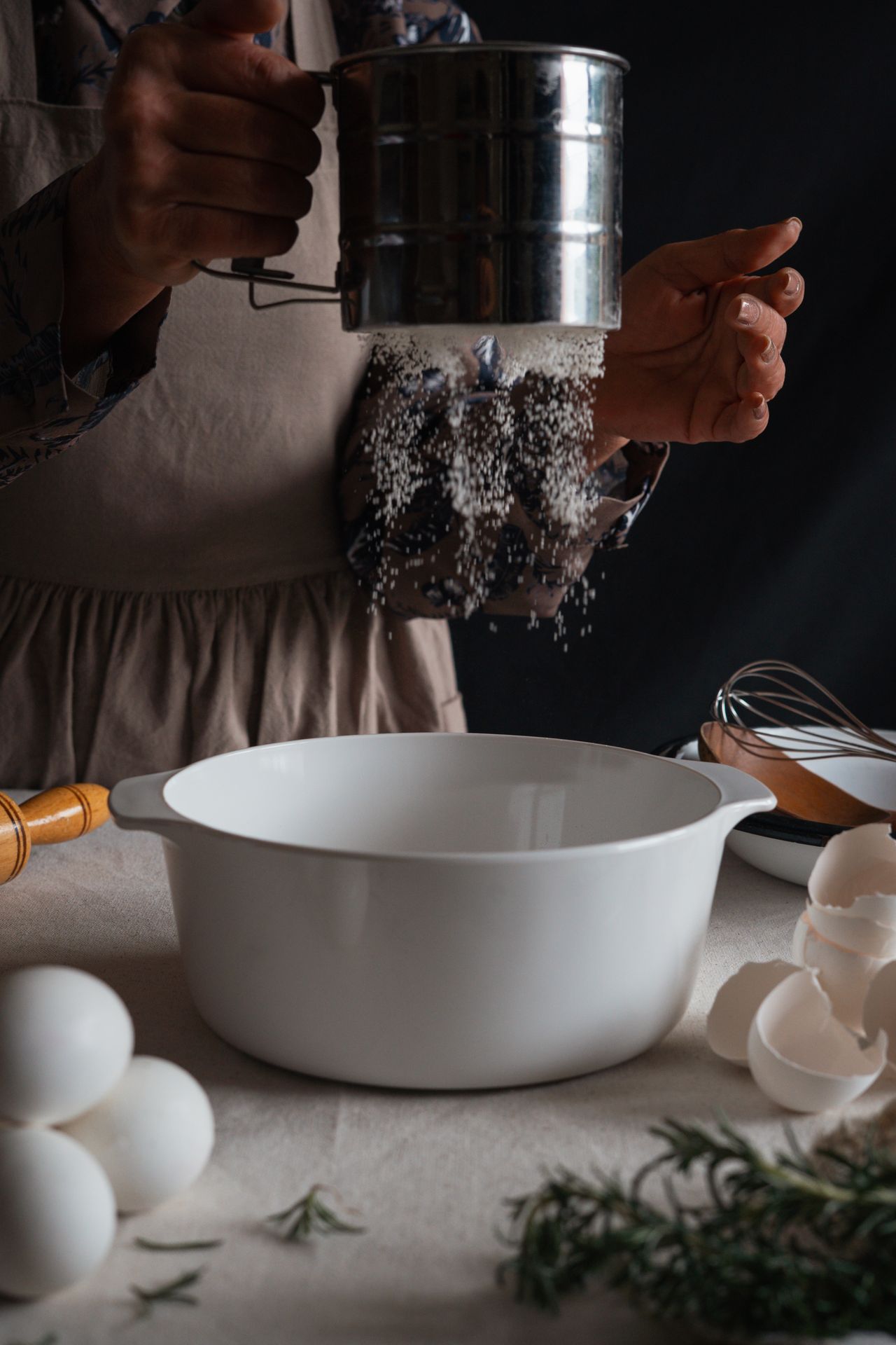 Flour falling from a metal sifter into a white bowl, with eggshells and herbs nearby.