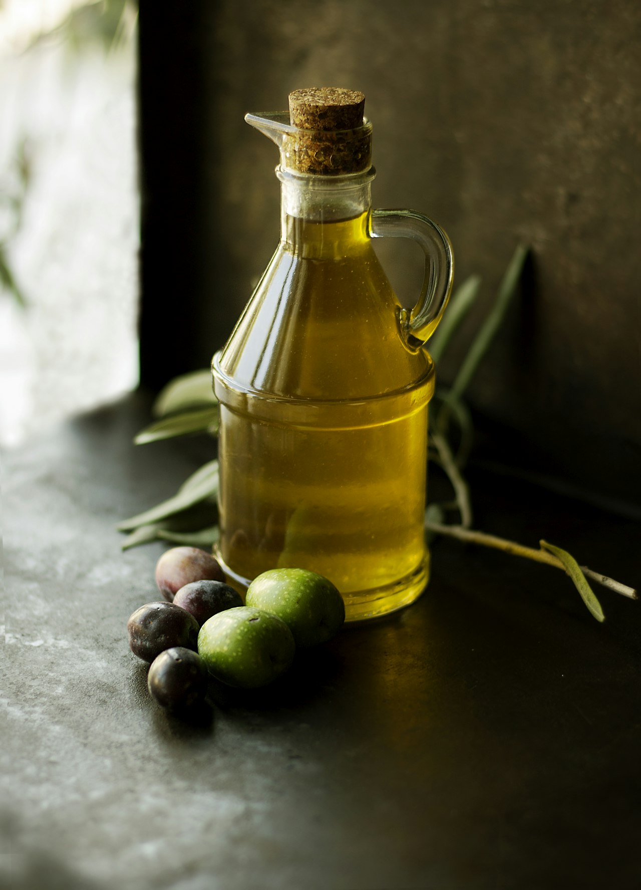 A glass cruet of olive oil stands beside olives and an olive branch.