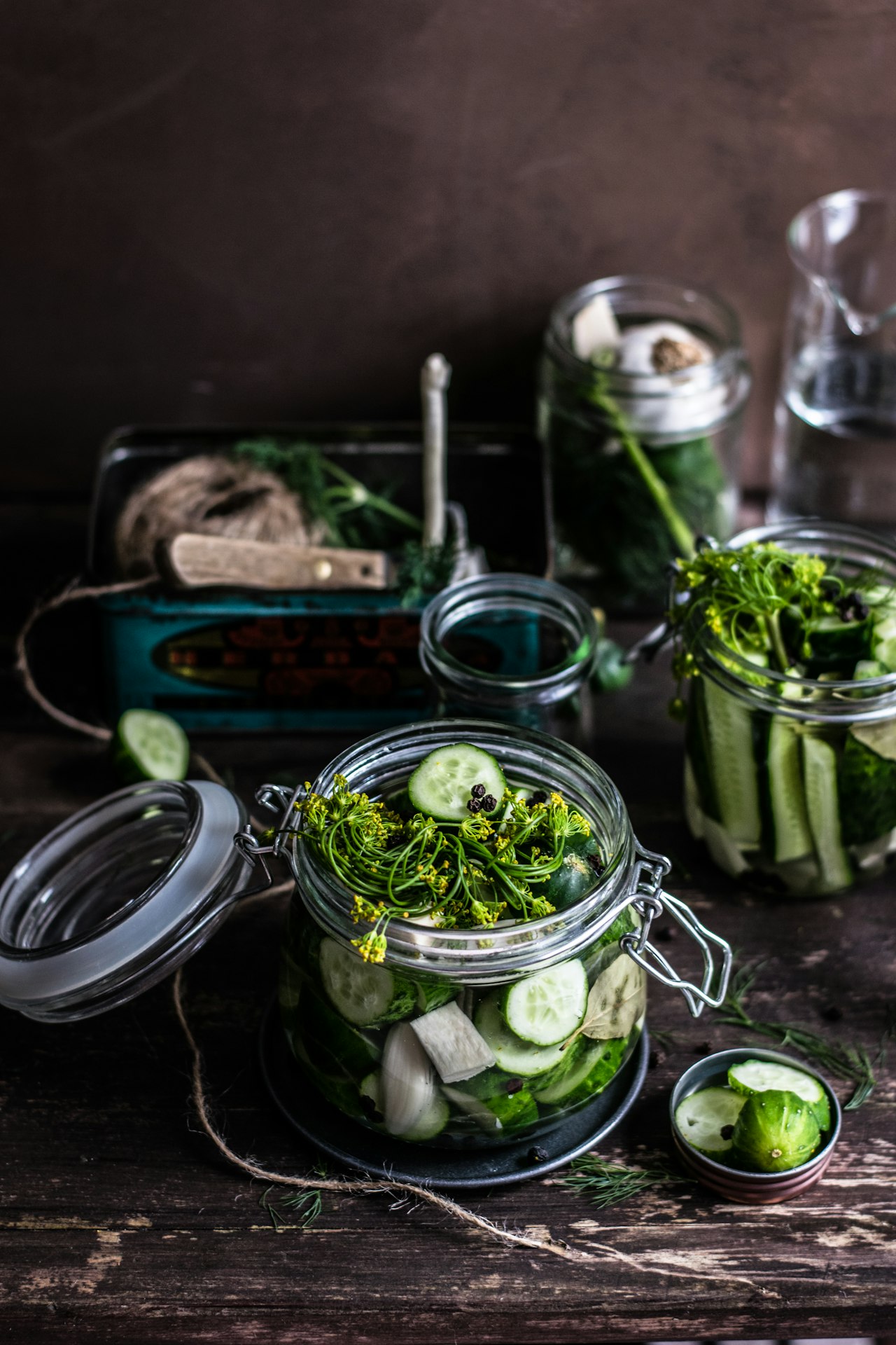 Open clip-top glass jars hold sliced cucumbers and herbs on a dark wooden table.