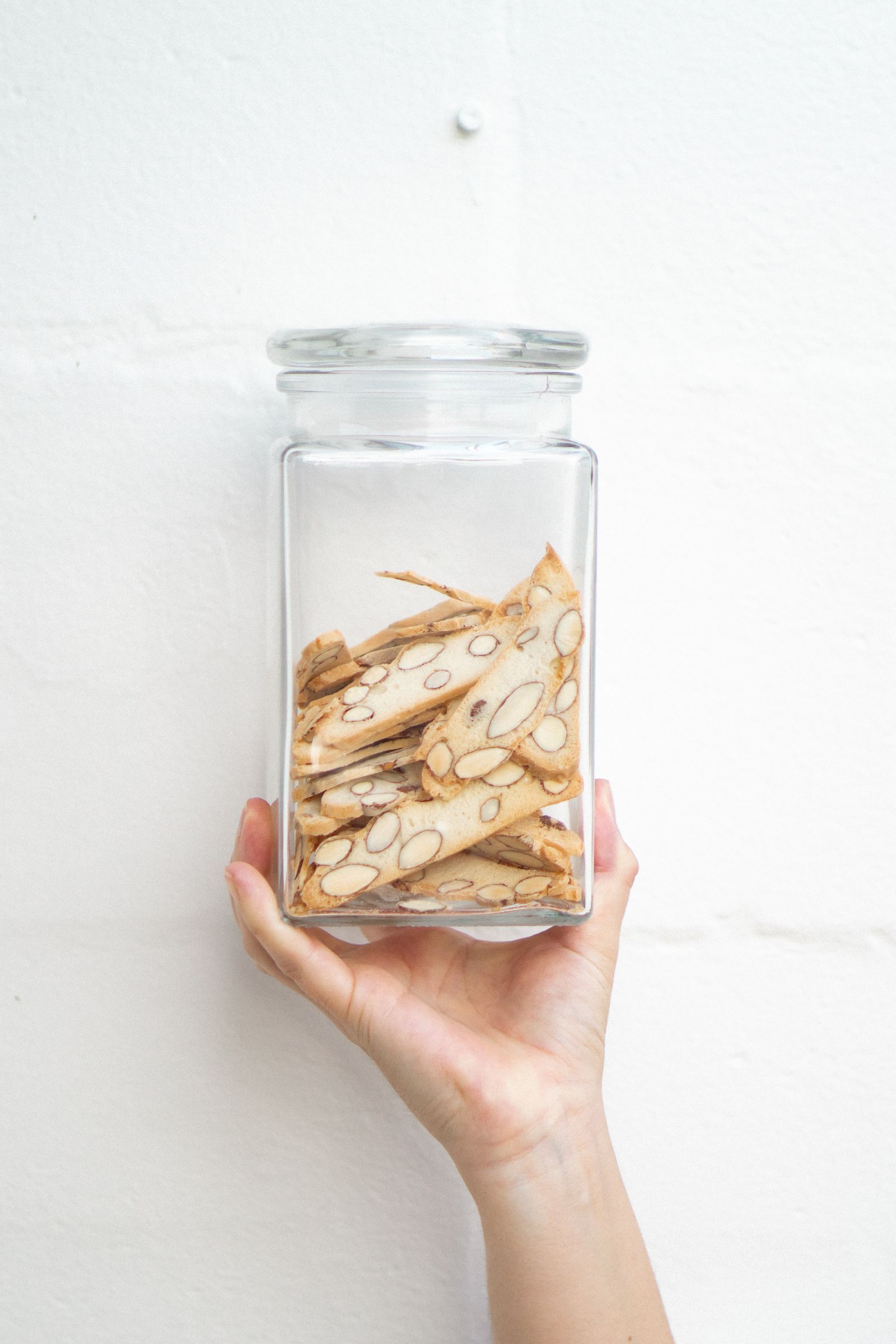 A hand holds a glass jar containing almond-studded biscotti against a white wall.