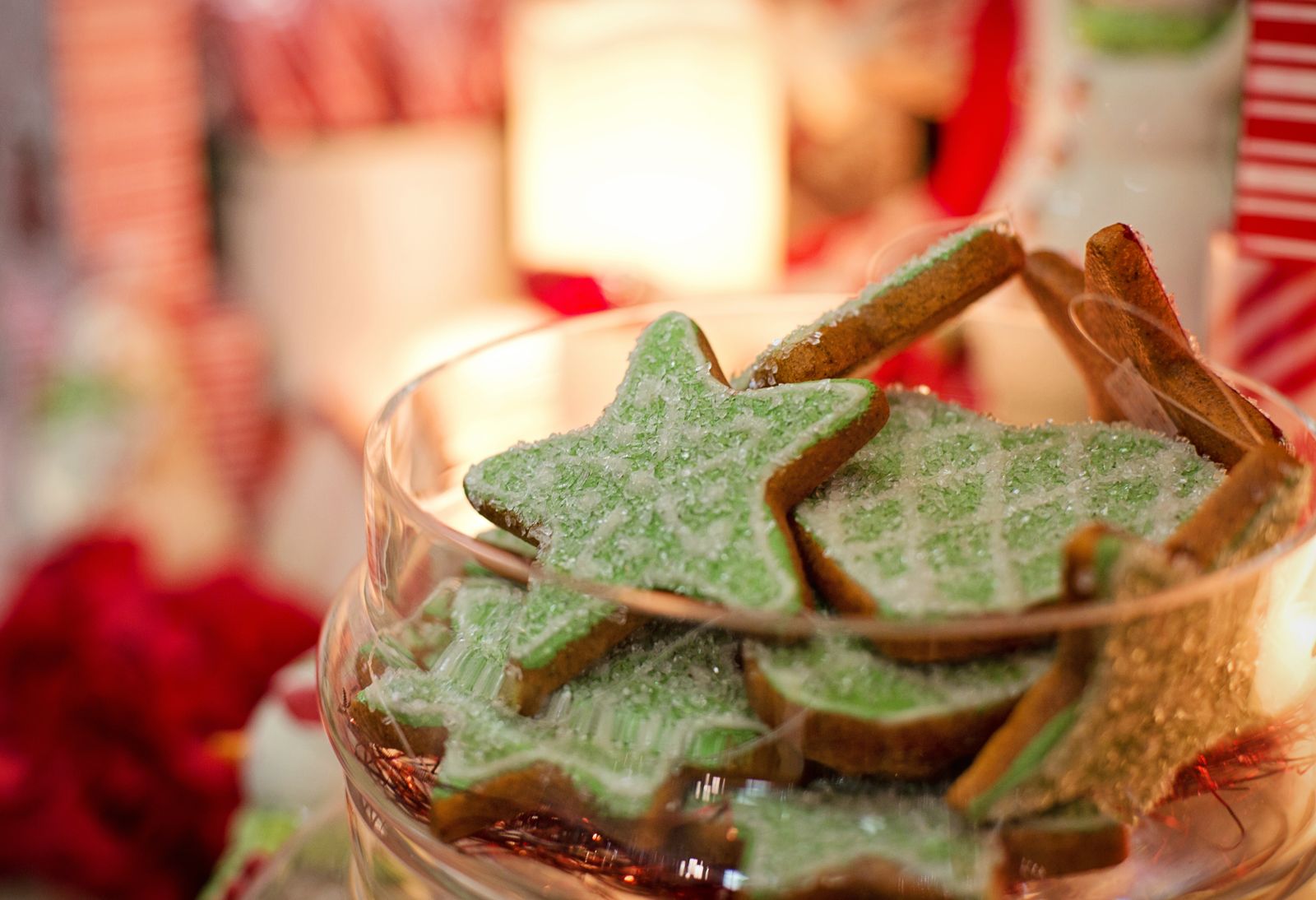 Green iced star cookies fill a glass container with blurred holiday decorations behind it.