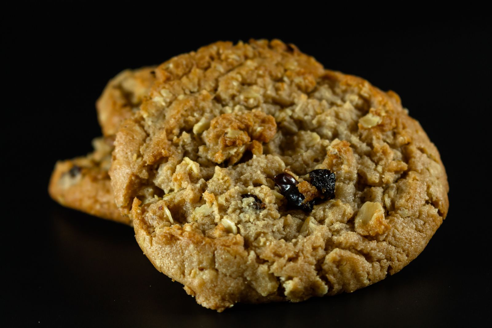 Oatmeal-raisin cookies overlap against a black background, with oat flakes and raisins visible.