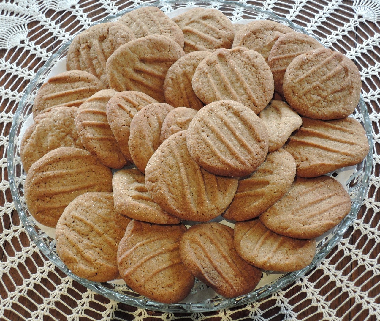 Round brown cookies with fork marks overlap on a glass plate over lace.
