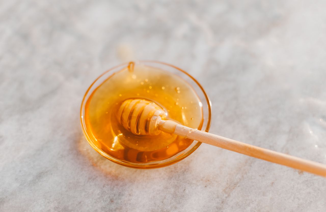 A wooden honey dipper rests across a glass bowl of honey on a pale counter.