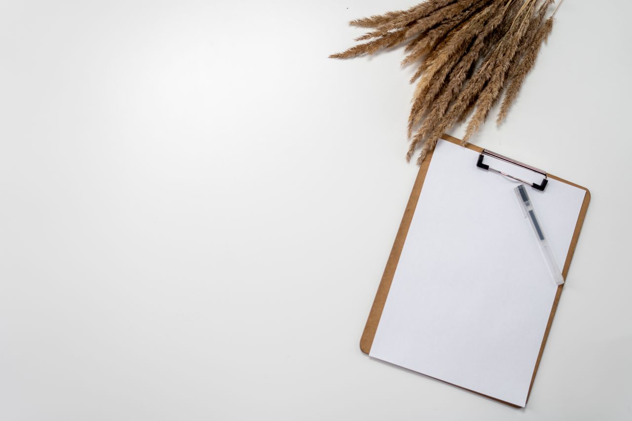 A blank clipboard and pen lie beside dried grasses on a white surface.