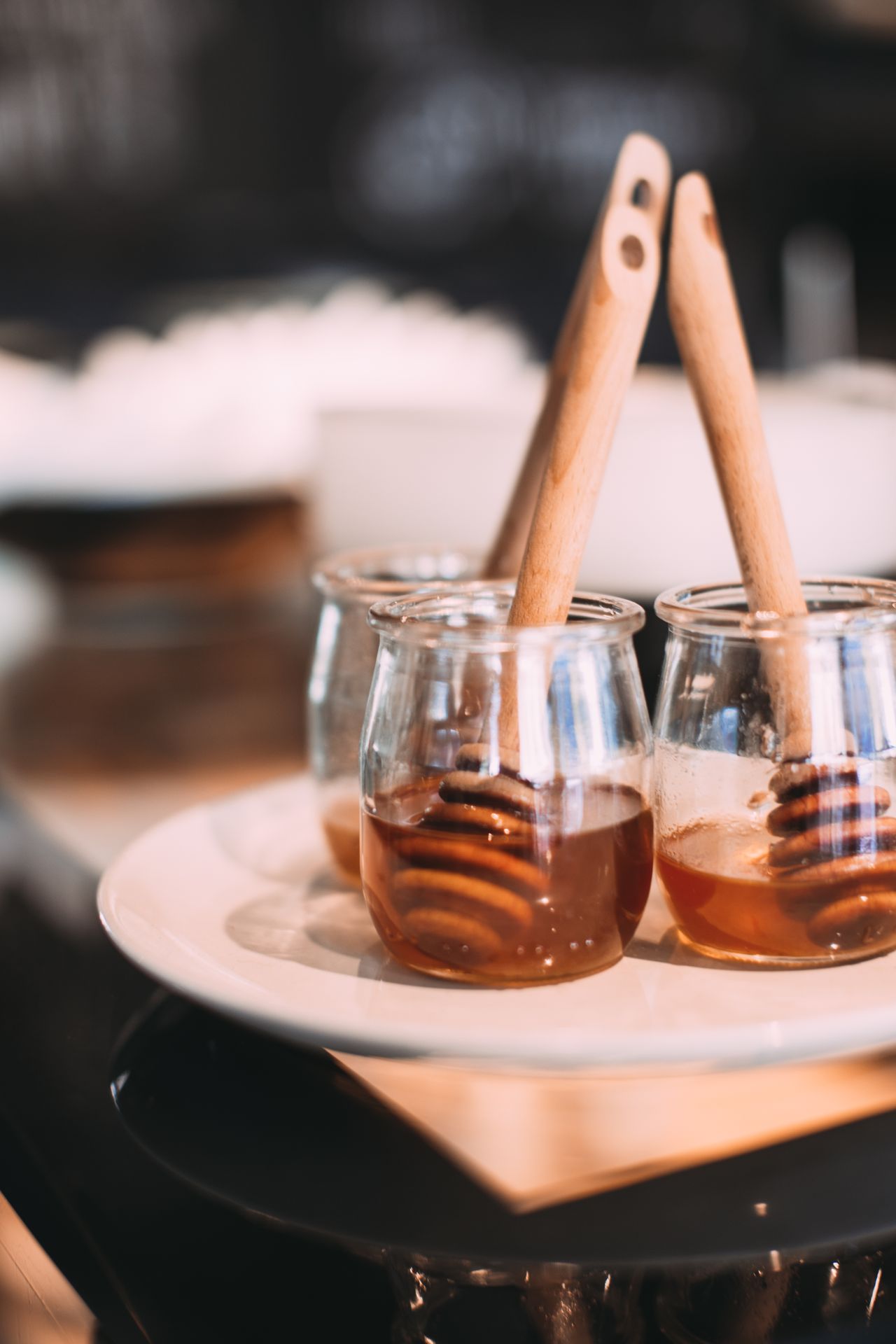 Three small glass jars of honey with wooden dippers sit on a pale plate.