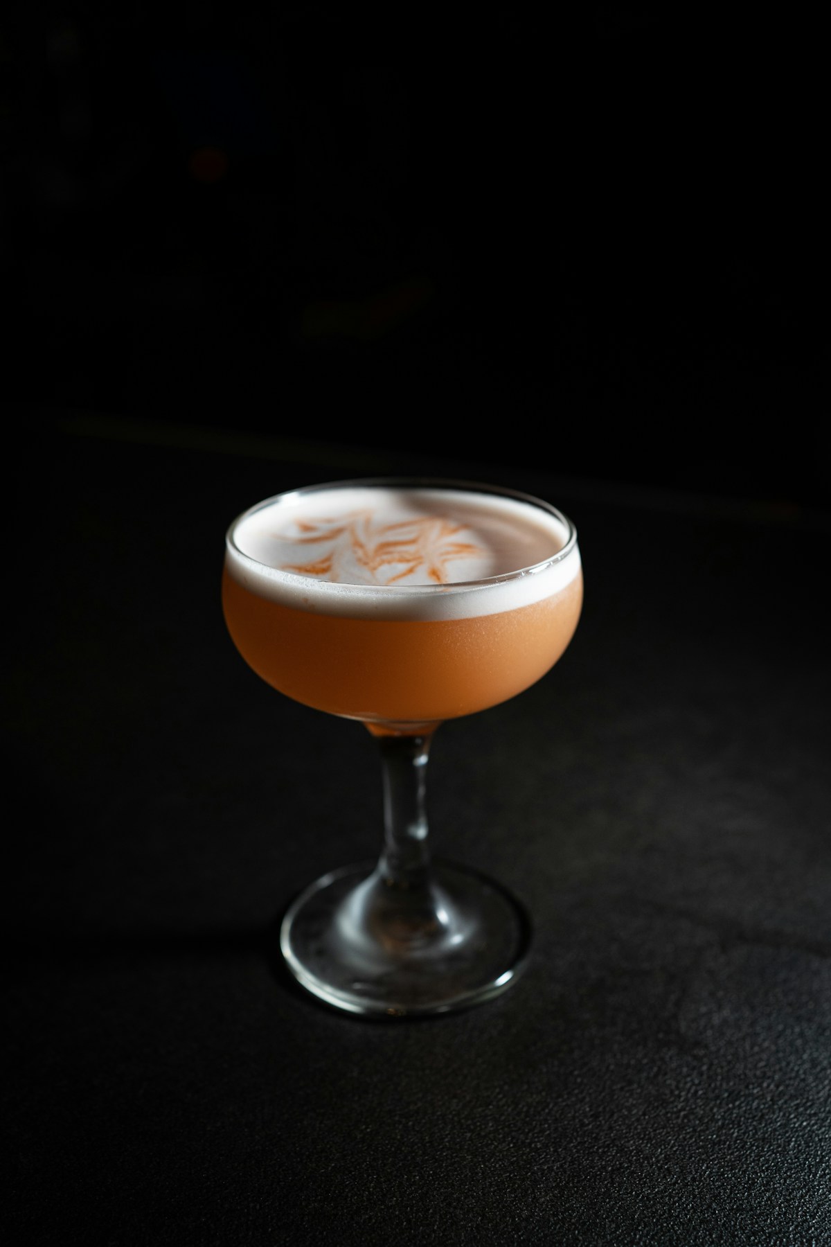 An orange-brown cocktail with a foamy top sits in a coupe glass on a dark surface.