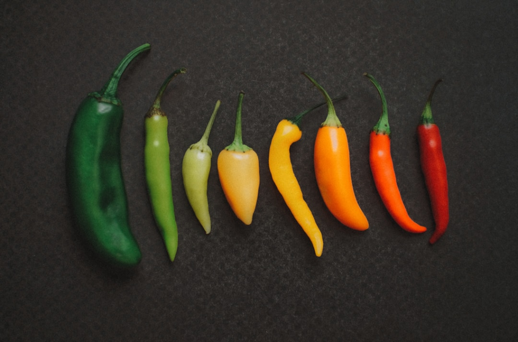 Eight green, yellow, orange and red peppers lie in a row on a dark surface.