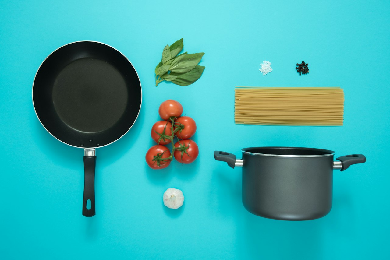A frying pan, pot, dry spaghetti, tomatoes, basil, garlic, salt and pepper sit on a turquoise surface.