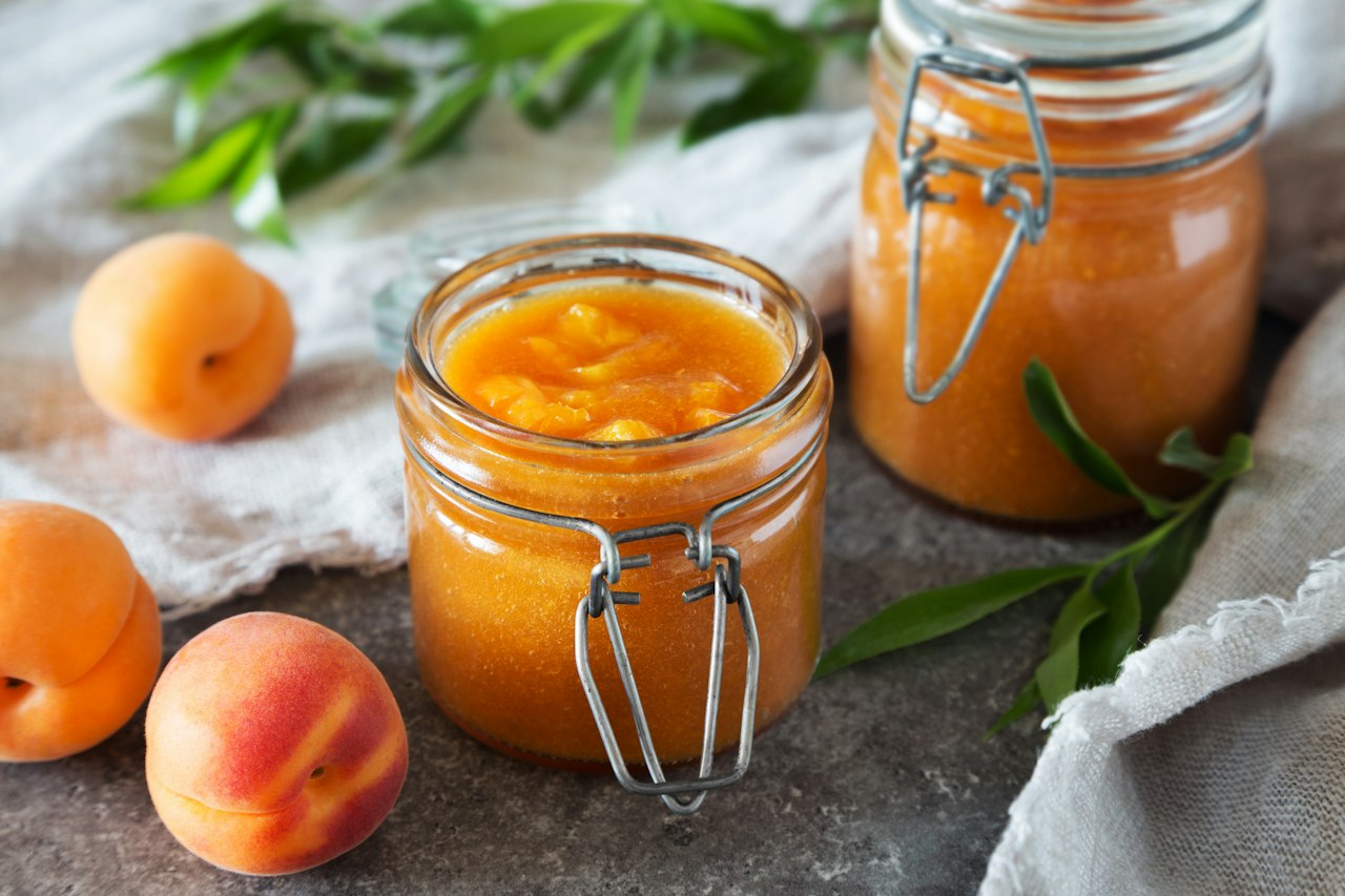 Two clamp-top jars hold an orange fruit mixture beside whole fruit and green leaves on gray cloth.