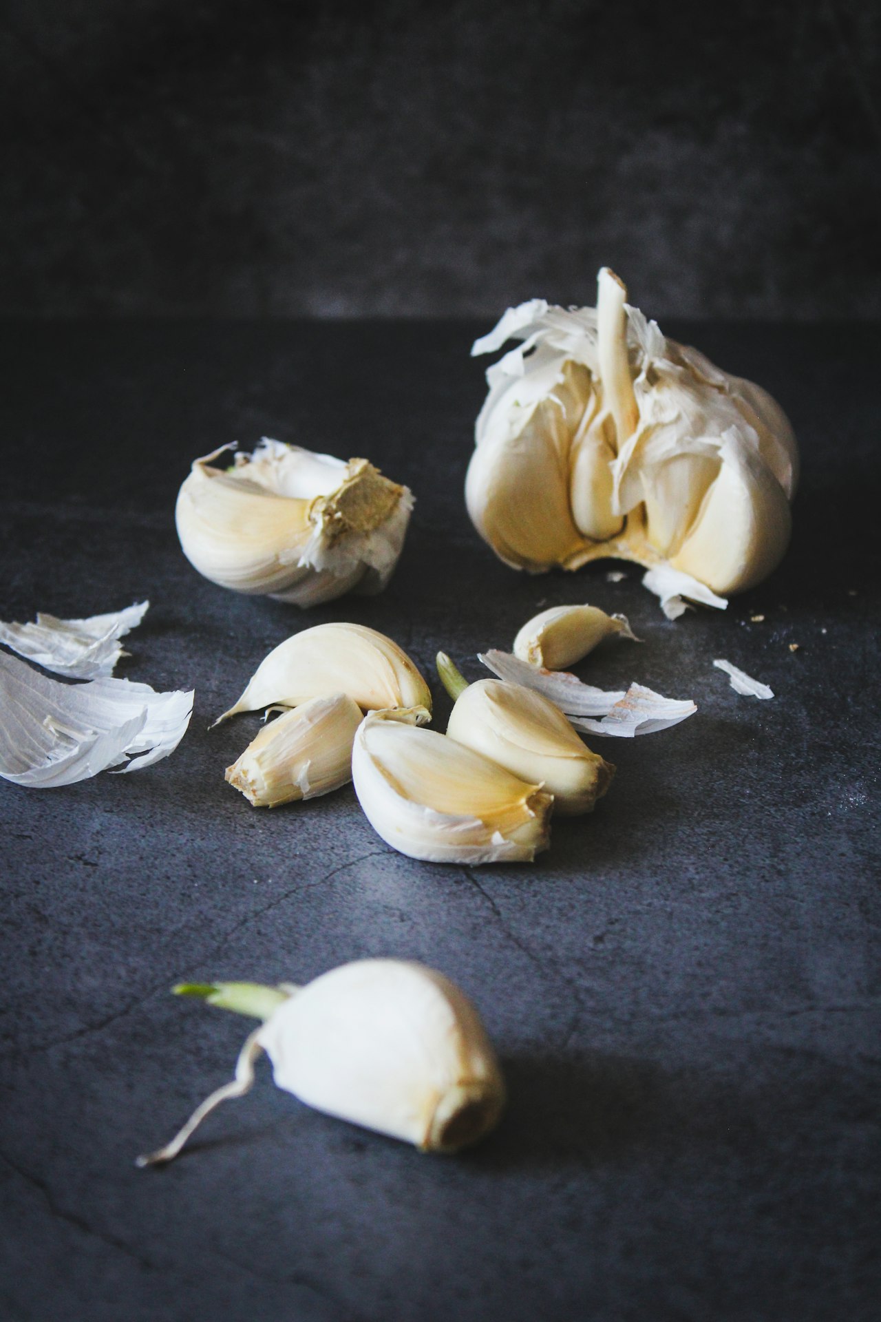 A broken garlic bulb, loose cloves and papery skins rest on a dark tabletop.