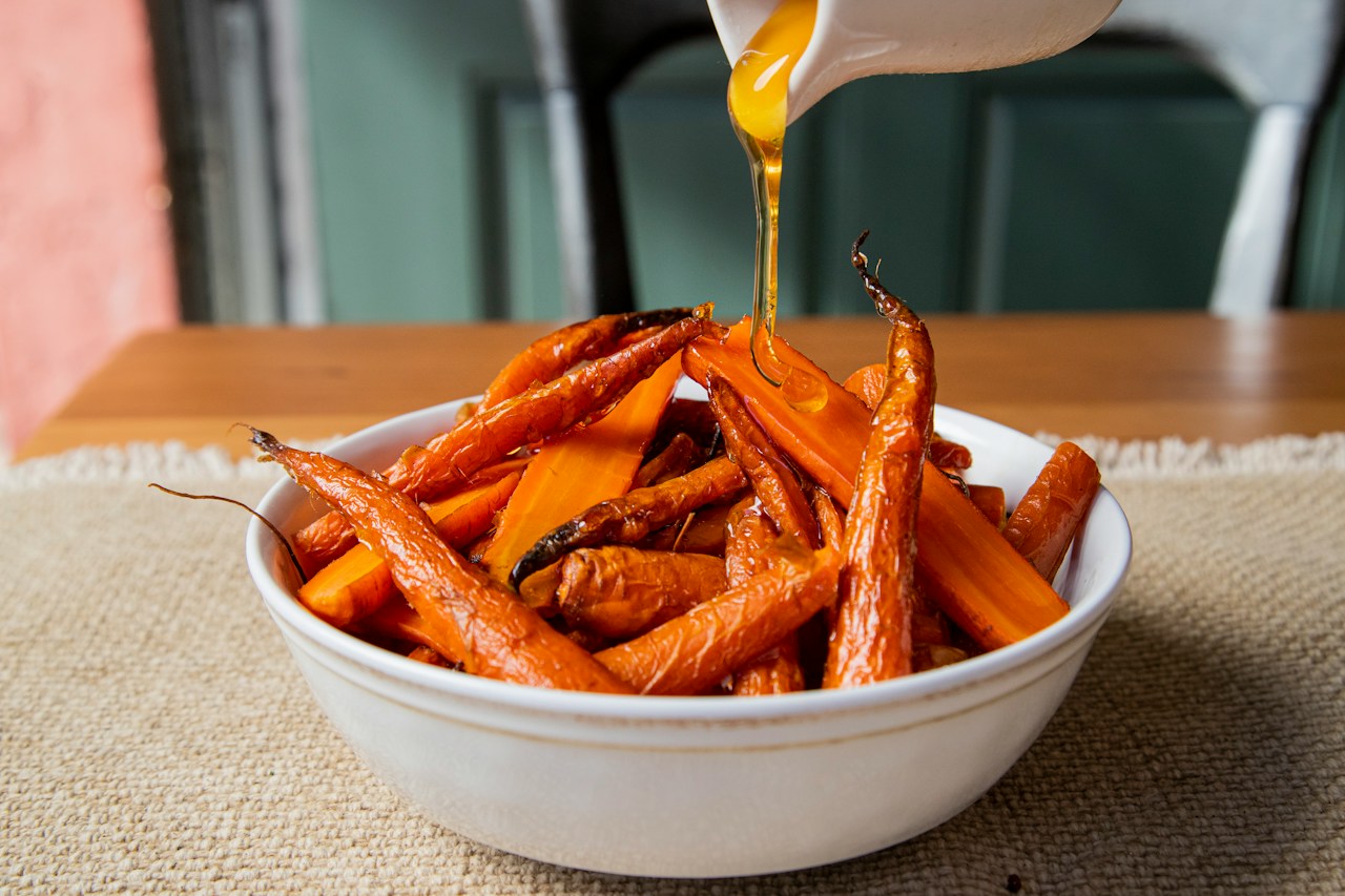 Golden liquid pours from a white pitcher onto roasted carrots in a white bowl.