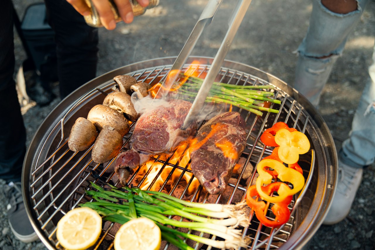 Meat, mushrooms, peppers and green vegetables cook on a round barbecue grill.