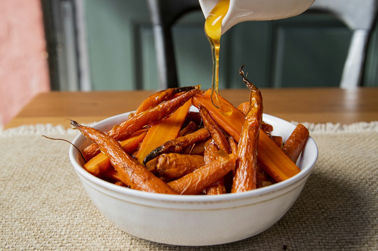 Amber liquid pours from a jug onto roasted carrots in a white bowl.