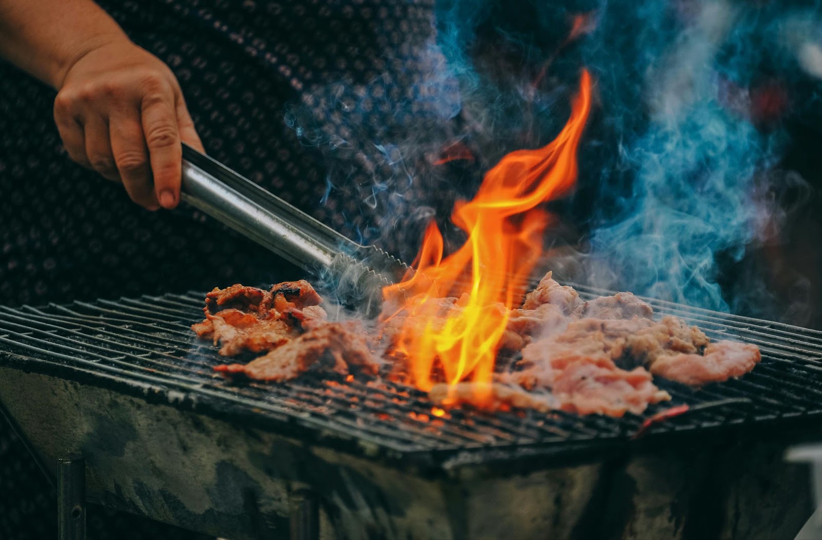 Tongs above meat on a smoky grill with flames.