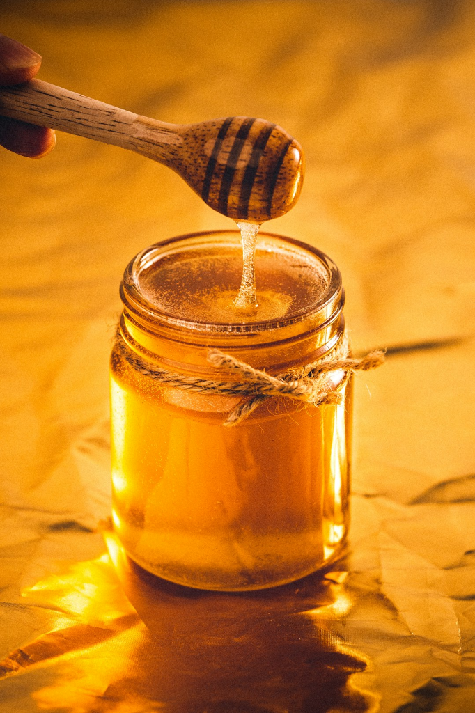A glass jar of honey and a honey dipper against a golden background.